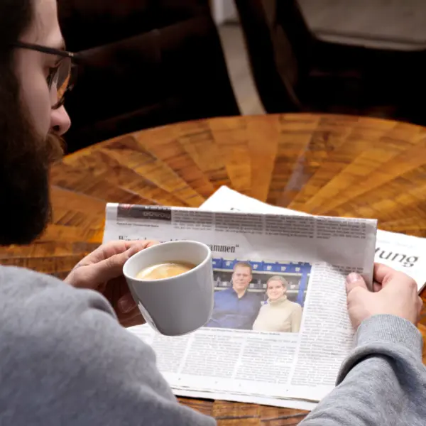 Person sitzt an einem runden Holztisch, hält eine Tasse Kaffee in der Hand und liest eine aufgeschlagene Zeitung mit einem Foto von zwei Personen.