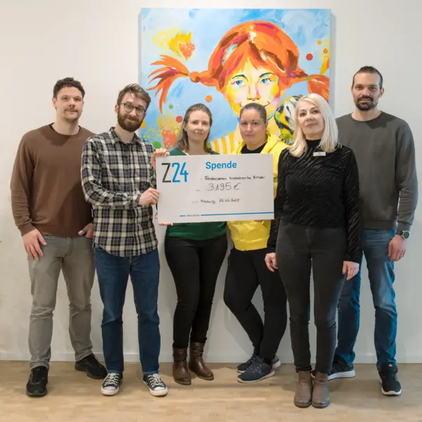 Gruppenfoto von sechs Personen, die einen symbolischen Spendenscheck der Firma Z24 über 3.195 Euro an den Förderverein krebskranker Kinder übergeben. Im Hintergrund hängt ein farbenfrohes Gemälde eines Kindes mit roten Zöpfen.