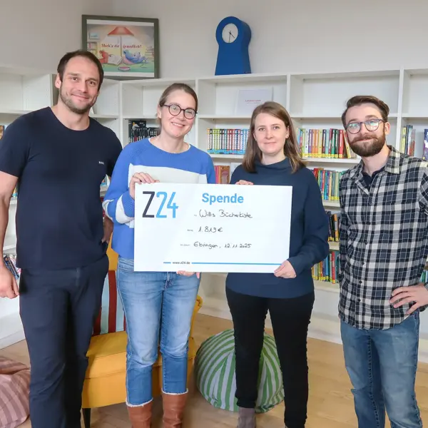 Vier Erwachsene stehen nebeneinander in einem hellen Raum mit Bücherregalen im Hintergrund. Zwei Personen in der Mitte halten einen großen Scheck mit der Aufschrift „Z24 Spende“ und einem Betrag. Alle lächeln in die Kamera.