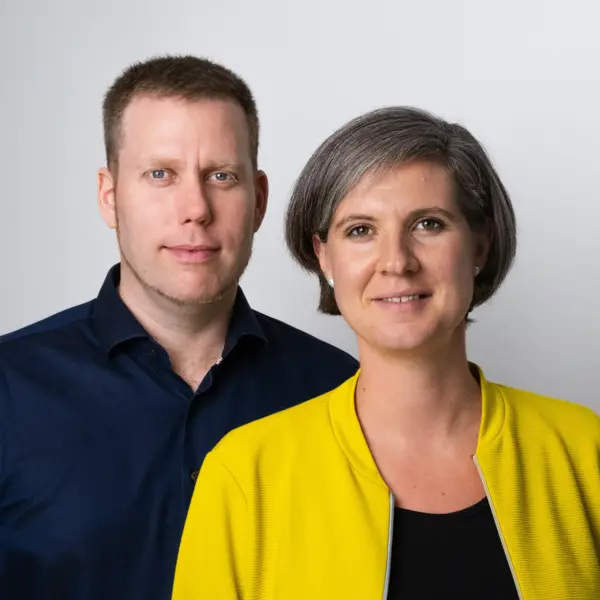 Michael Ebner-Seibold und Tabea Seibold stehen nebeneinander vor einem neutralen, hellgrauen Hintergrund. Tabea Seibold rechts trägt eine gelbe Jacke über einem schwarzen Oberteil und hat kurzes graues Haar. Michael Ebner-Seibold links trägt ein dunkelblaues Hemd und hat kurzes, hellbraunes Haar. Beide blicken freundlich in die Kamera.
