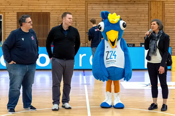 Im Vordergrund stehen in einer Reihe ein Mann von den Eisvögeln, Herr Ebner-Seibold, das Maskottchen Birdie und Frau Seibold. Frau Seibold hält ein Mikrofon in der Hand und hält eine kleine Rede.