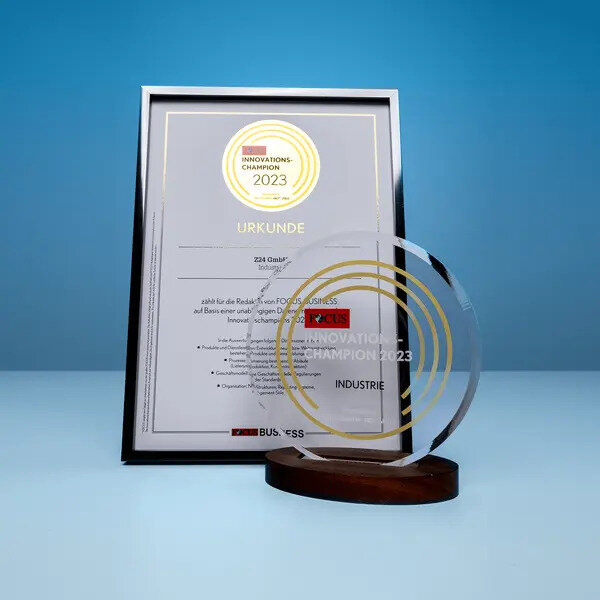 Gerahmte Urkunde und transparente Trophäe mit der Aufschrift "FOCUS Innovationschampion 2023" auf einem Tisch, vor blauem Hintergrund.