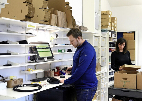 Ein Mann und eine Frau arbeiten in einem Lager; der Mann scannt Produkte an einem Computer, die Frau packt Kartons aus. Regale mit Kartons und Verpackungsmaterialien im Hintergrund.