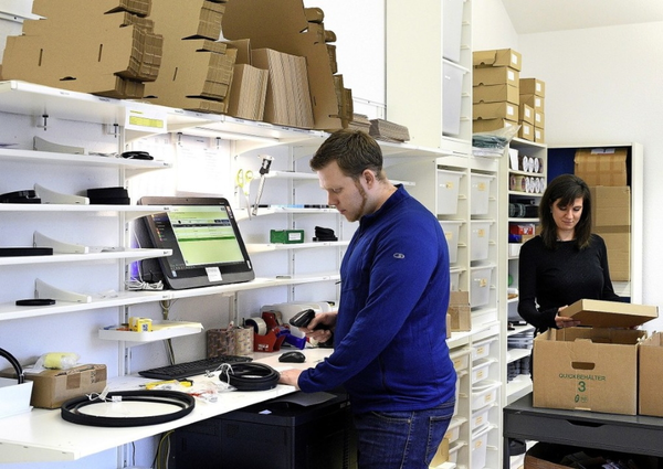 Ein Mann und eine Frau arbeiten in einem Lager; der Mann scannt Produkte an einem Computer, die Frau packt Kartons aus. Regale mit Kartons und Verpackungsmaterialien im Hintergrund.