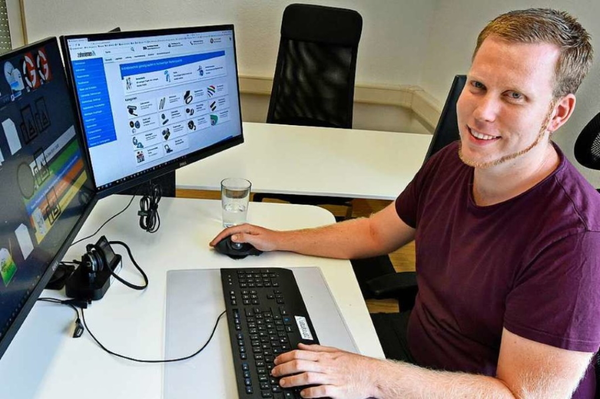 Mann vor zwei Monitoren, auf denen eine Website mit technischen Produkten angezeigt wird; er sitzt an einem Schreibtisch mit Tastatur und Glas Wasser.