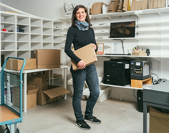 Eine Frau in einem Lagerraum hält ein Paket; im Hintergrund sind Regale mit Kartons, ein Drucker und Bürozubehör sichtbar.