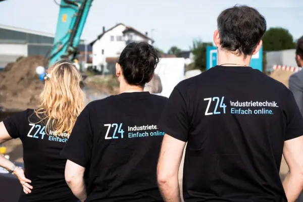 Zwei Frauen und ein Mann stehen mit dem Rücken zum Bild. Auf den schwarzen T-Shirts, die sie tragen, ist das Logo von Z24 zu sehen mit dem Slogan: "Industrietechnik. Einfach online.". Sie schauen auf die Baustelle, auf der ein Bagger steht.