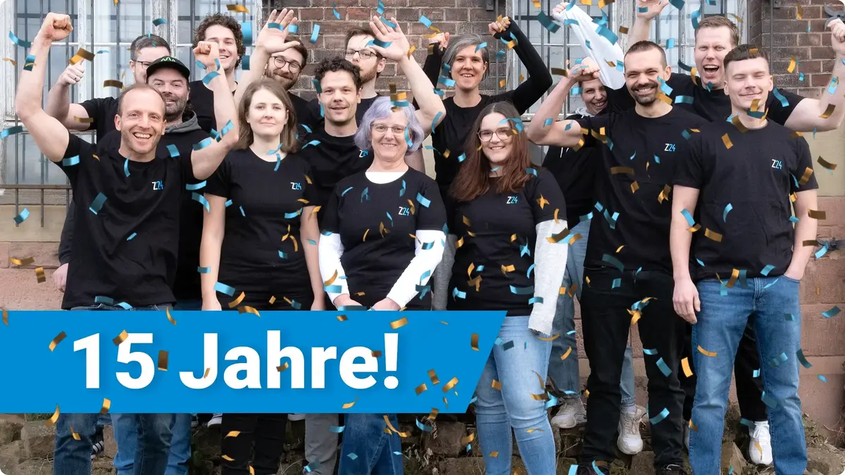 Eine Gruppe der Mitarbeitenden von Z24 jubelt gesammelt im einheitlichen Z24 T-Shirt vor einem älteren Gebäude. Im Vordergrund die Aufschrift in weiß auf blauem Hintergrund "15 Jahre!"