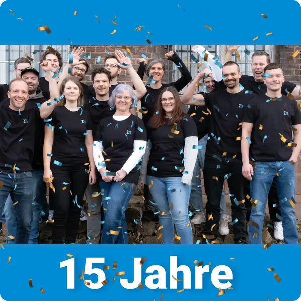 Eine Gruppe der Mitarbeitenden von Z24 jubelt gesammelt im einheitlichen Z24 T-Shirt vor einem älteren Gebäude. Oben und unten befindet sich ein blauer Balken mit der Aufschrift unten in weiß "15 Jahre".