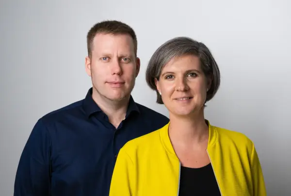 Michael Ebner-Seibold und Tabea Seibold stehen nebeneinander vor einem neutralen, hellgrauen Hintergrund. Tabea Seibold rechts trägt eine gelbe Jacke über einem schwarzen Oberteil und hat kurzes graues Haar. Michael Ebner-Seibold links trägt ein dunkelblaues Hemd und hat kurzes, hellbraunes Haar. Beide blicken freundlich in die Kamera.