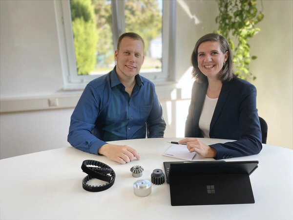 Zwei Personen sitzen lächelnd an einem runden Tisch. Vor ihnen liegen verschiedene technische Bauteile und ein Laptop. Im Hintergrund ist ein Fenster mit Pflanzen zu sehen.