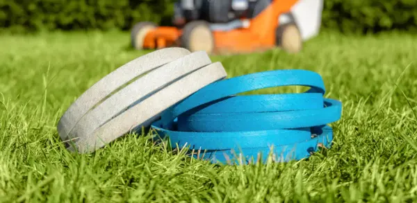 Zwei Keilriemen liegen im Gras vor einem unscharfen orangefarbenen Rasenmäher.