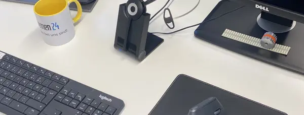 Tisch mit Computerzubehör, darunter eine Tastatur, Maus, Monitor und Headsethalterung. Eine Tasse mit gelbem Rand, auf der das Firmenlogo zu erkennen ist und eine kleine Zahnriemenscheibe auf einem Zahnriemen sind ebenfalls zu sehen.