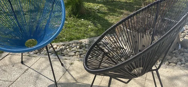 Zwei moderne, bogenförmige Stühle, einer in Blau und der andere in Schwarz, stehen auf einer Terrasse mit Steinboden. Im Hintergrund sind Rasen und Kieselsteine sichtbar.