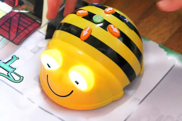 Ein faustgroßer Bienenroboter mit leuchtenden Augen und einen runden, grünen Knopf in der Mitte, welcher von weißen Pfeilen auf rotem Hintergrund umgeben ist, fährt auf weißem Papier hin und her.