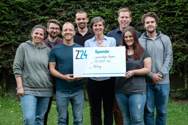 Acht Personen stehen draußen in einer Reihe und halten gemeinsam einen großen Spendenscheck über 10.000 Euro.