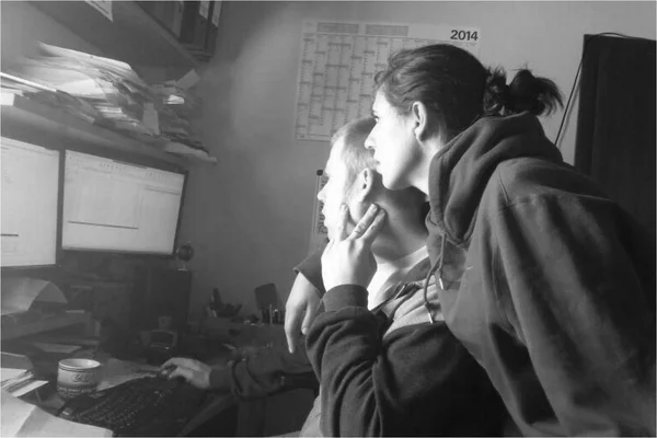 Ein Mann und eine Frau schauen gemeinsam auf einen Computerbildschirm in einem Büro, umgeben von Papieren und Ordnern; eine Jahreszahl 2014 ist an der Wand sichtbar.