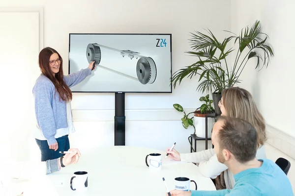 Eine Frau in einem hellen Seminarraum präsentiert einem kleinen Team eine technische Zeichnung auf einem Bildschirm. Die Teammitglieder sitzen an einem Tisch und machen sich Notizen. Im Hintergrund steht eine Pflanze.
