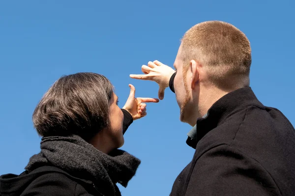 Zwei Personen in schwarzer Kleidung formen einen rechteckigen Rahmen mit ihren Händen und blicken in den Himmel.