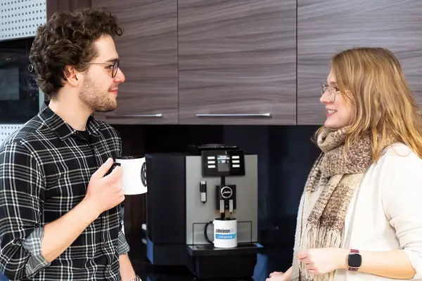 Ein Mann und eine Frau unterhalten sich lächelnd vor einer Kaffeemaschine, beide halten Tassen in der Hand.