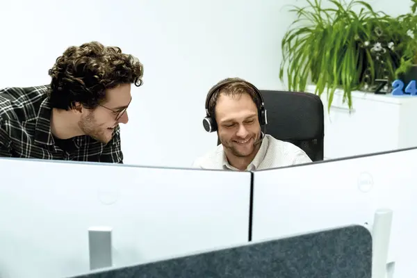 Zwei Männer arbeiten lächelnd an Computern, einer trägt Kopfhörer. Im Hintergrund sind grüne Pflanzen sichtbar.