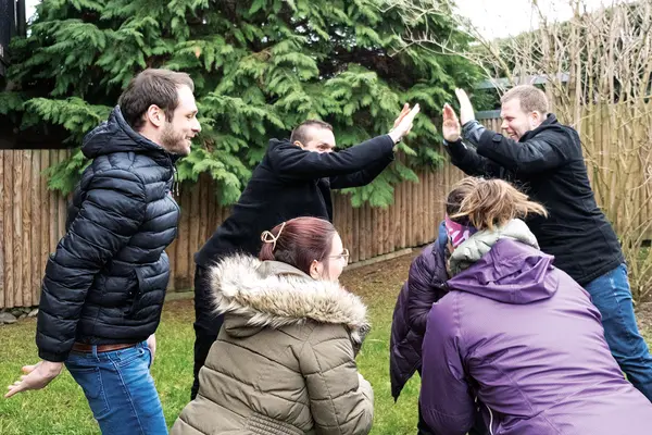 Personengruppe, die im Freien eine Teamaktivität macht, mit zwei Personen, die sich abklatschen. Sie tragen Winterkleidung und stehen auf einer Wiese vor dichtem Nadelbaumbewuchs.