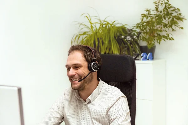 Ein Mann mit Headset sitzt lächelnd in einem Bürostuhl vor einem Computerbildschirm, im Hintergrund stehen Pflanzen und eine Dekoration mit der Zahl 24.