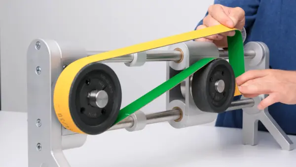 Demonstrationsaufbau mit zwei Umlenkrollen, über die zwei flache Antriebsriemen in Gelb und Grün geführt werden. Eine Person spannt die Riemen mit beiden Händen, um die Vorspannung zu zeigen