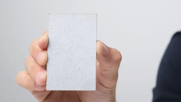 Rechteckiges, hellgraues Materialmuster mit feinporiger Oberfläche wird zwischen Daumen und Fingern gehalten.