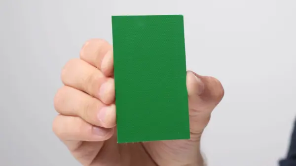 Rechteckiges, grünes Materialmuster mit glatter Oberfläche wird zwischen Daumen und Fingern gehalten.
