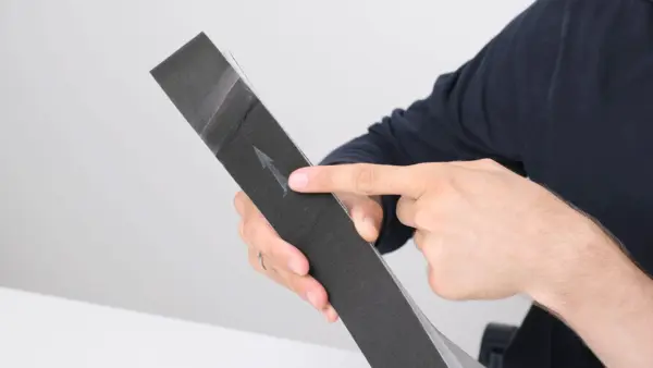 Flacher schwarzer Antriebsriemen wird schräg in der Hand gehalten; ein Finger zeigt auf einen aufgedruckten Pfeil, der die Laufrichtung des Riemens kennzeichnet.