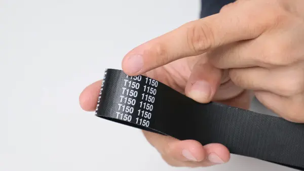 Nahaufnahme eines schwarzen Zahnriemens, der in der Hand gehalten wird. Ein Finger zeigt auf die weiße Riemenkennzeichnung ‚T150 1150‘ zur Identifikation von Teilung und Länge.