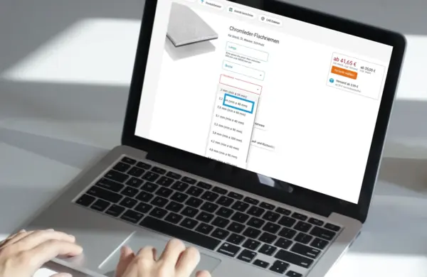 erson bedient einen Laptop, auf dessen Bildschirm eine Produktseite für Chromleder-Flachriemen mit Konfigurationsoptionen und Preisangaben angezeigt wird.