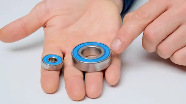 Zwei Kugellager mit blauer Dichtung liegen auf einer geöffneten Hand: links ein kleines Lager, rechts ein größeres. Ein Finger zeigt auf das größere Kugellager.