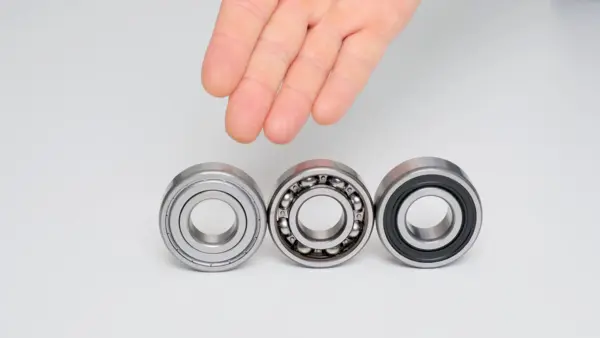 Drei nebeneinander stehende Kugellager auf hellem Hintergrund: links ein geschlossenes Metalllager, in der Mitte ein offenes Kugellager mit sichtbaren Kugeln, rechts ein Kugellager mit schwarzer Dichtung. Darüber schwebt eine menschliche Hand, als würde sie die Lager vergleichen oder auswählen.