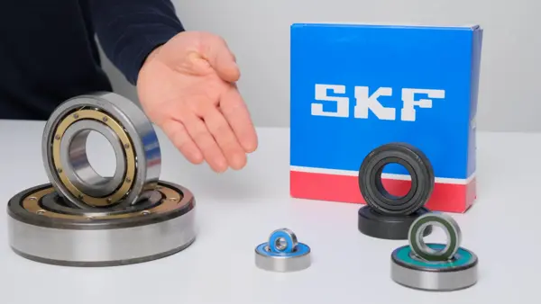 Verschiedene Kugellager liegen auf einem weißen Tisch: links zwei große, offene Metallkugellager übereinander, daneben eine ausgestreckte menschliche Hand. Rechts steht eine blaue SKF-Verpackung, davor mehrere kleinere Kugellager in unterschiedlichen Ausführungen.