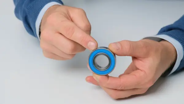 Kleines Kugellager mit blauer Dichtung wird zwischen beiden Händen gehalten, während ein Finger auf die Dichtung am Außenring zeigt.