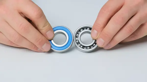Zwei Kugellager werden nebeneinander gehalten: links ein Kugellager mit blauer Dichtung, rechts ein offenes Kugellager mit sichtbaren Stahlkugeln, zum direkten Vergleich.