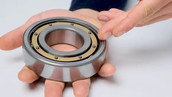 Großes, offenes Kugellager wird in einer geöffneten Hand gehalten, während ein Finger auf den äußeren Lagerring zeigt; der Käfig und die Kugeln sind sichtbar.