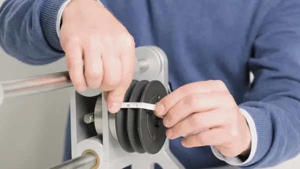 Eine Person misst mit einem Maßband den Durchmesser einer schwarzen Riemenscheibe, die an einer Maschine montiert ist.