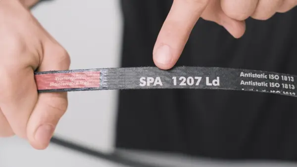 Eine Person hält einen schwarzen Keilriemen in den Händen, während ein Finger auf die weiße Aufschrift ‚SPA 1207 Ld‘ zeigt, die die Riemenspezifikation kennzeichnet.