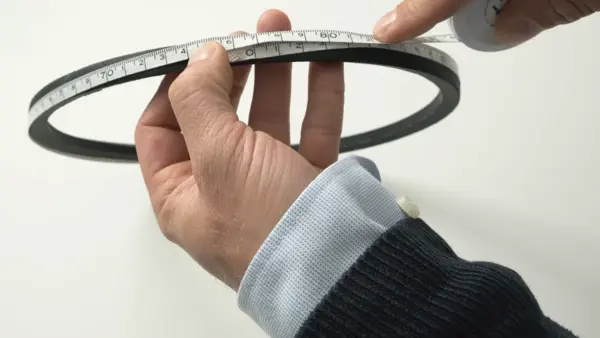 Eine Person misst mit einem Maßband den Umfang eines schwarzen Keilriemens, den sie in den Händen hält.