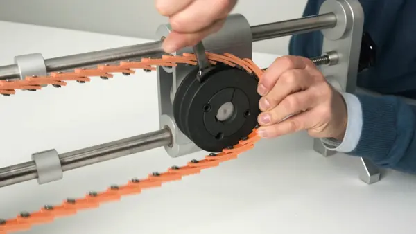 Person montiert einen orangefarbenen Keilriemen mit mechanischen Verbindern auf eine schwarze Riemenscheibe in einer Spannvorrichtung.