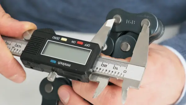 Eine Hand hält eine Industriekette mit der Prägung "16B", während mit einer digitalen Schieblehre das Maß eines Kettenglieds überprüft wird.