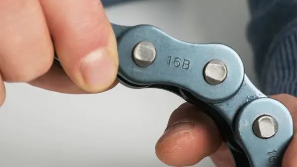 Nahaufnahme einer Industriekette, die von zwei Händen gehalten wird. Auf dem Kettenglied ist die Prägung "16B" zu sehen.