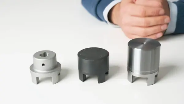 Drei unterschiedliche Kupplungsnaben aus Metall und Kunststoff sind nebeneinander aufgereiht, während eine Person im Hintergrund sitzt und die Hände auf dem Tisch hält.