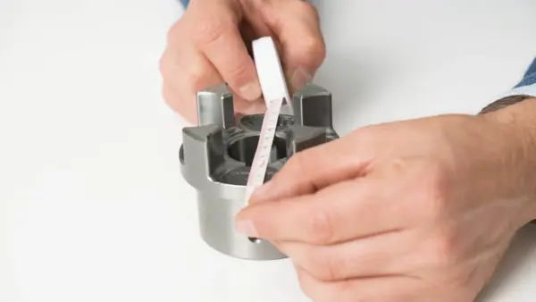 Eine Person misst mit einem Maßband den Innendurchmesser einer Metallkupplung, die auf einem weißen Tisch liegt.