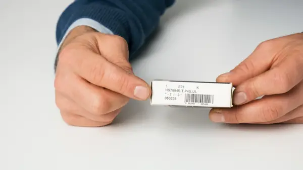 Hände zeigen eine weiße Verpackung mit aufgedruckter Lagernummer und Barcode.