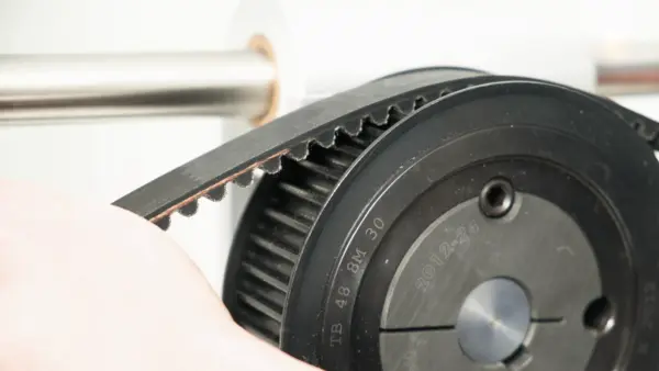 Nahaufnahme einer schwarzen Zahnriemenscheibe, über die ein Zahnriemen gespannt wird. Die Riemenscheibe ist an einer Metallwelle montiert, Beschriftungen wie ‚TB 48 8M 30‘ sind auf der Scheibe sichtbar.