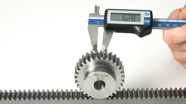 Eine Person misst mit einer digitalen Schieblehre den Teilkreisdurchmesser eines Zahnrads, das mit einer Zahnstange im Eingriff steht. Auf der Anzeige der Schieblehre ist der Wert 6,28 mm zu sehen